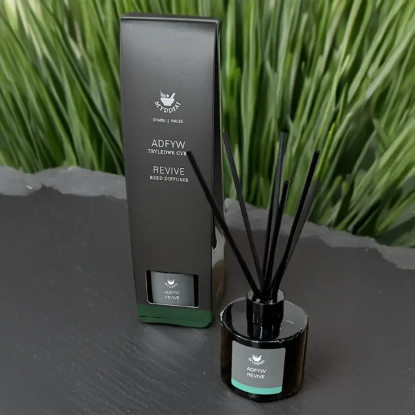 100ml glass reed diffuser bottle labelled “Revive”, accompanied by natural reeds and presented on a clean surface. The design is minimalist and elegant, with a subtle Welsh-made branding visible.